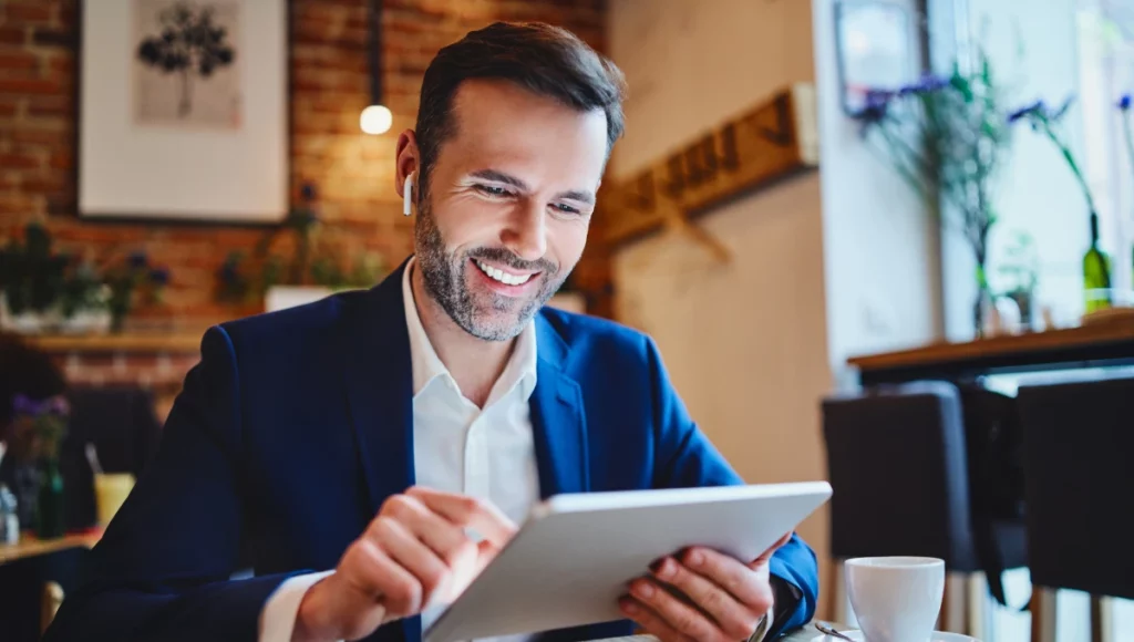 Businessman sitting in cafe looking at tablet during phone call through wireless headphones